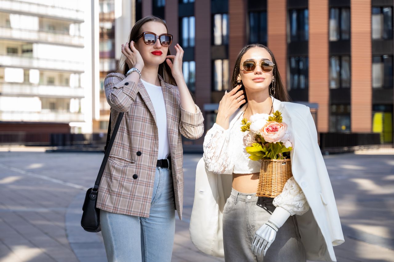 About Two stylish women enjoy a sunny day in the city wearing chic outfits and sunglasses.